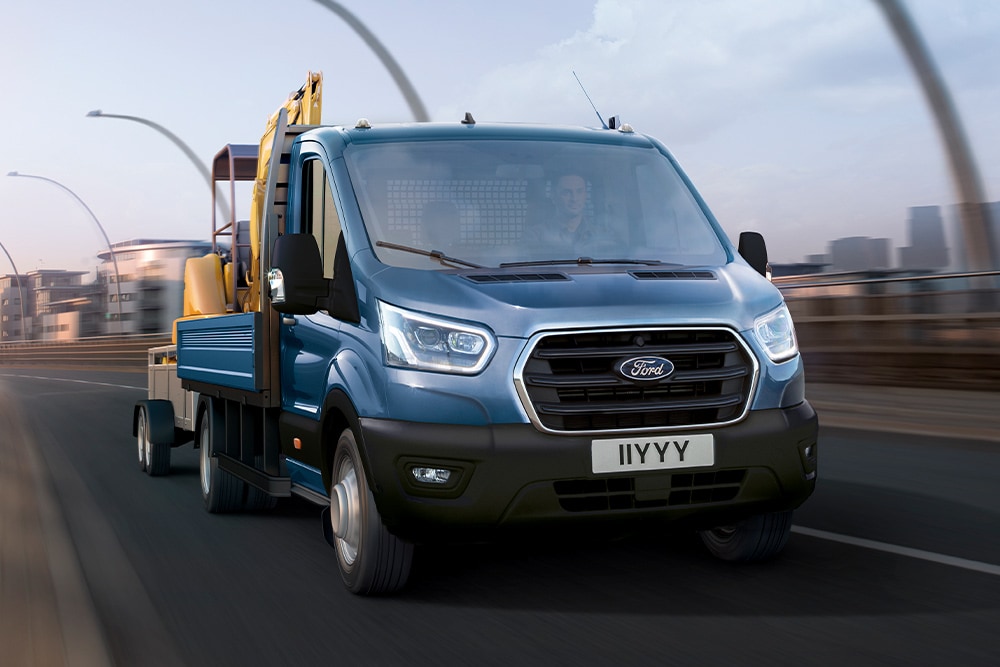Frontansicht eines Ford Transit Fahrgestells mit geladenem Bagger bei der Fahrt durch die Stadt.