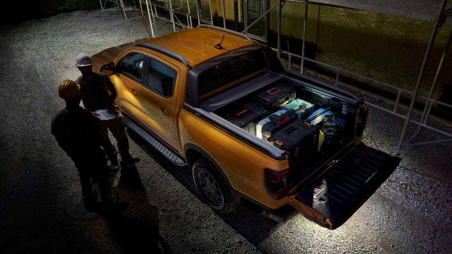 Ford Ranger in Orange in der Vogelperspektive mit beladener Ladefläche parkt bei Dunkelheit vor Wohnhausneubau neben zwei Personen