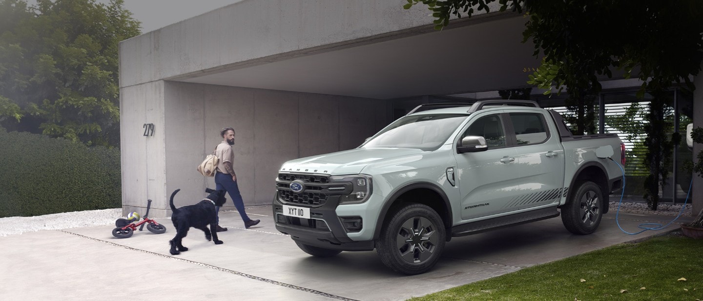 Ein Ford Ranger Plug-in-Hybrid ist in einer Einfahrt geparkt und an eine Ladestation angeschlossen. Ein Mann geht mit einem schwarzen Hund auf das Fahrzeug zu.