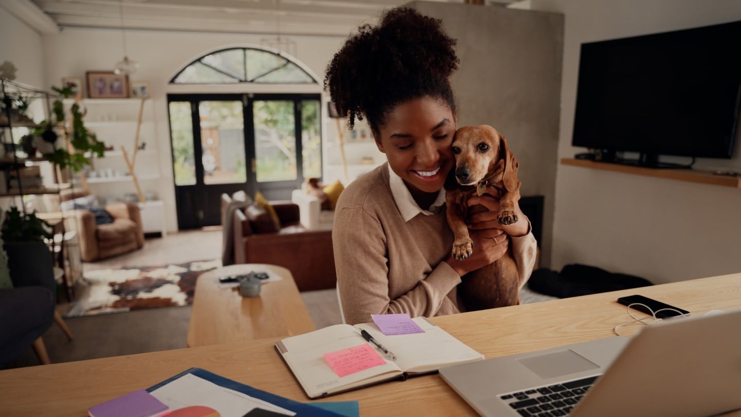 Frau in ihrem Zuhause mit Hund auf dem Arm lächelnd vor einem Laptop