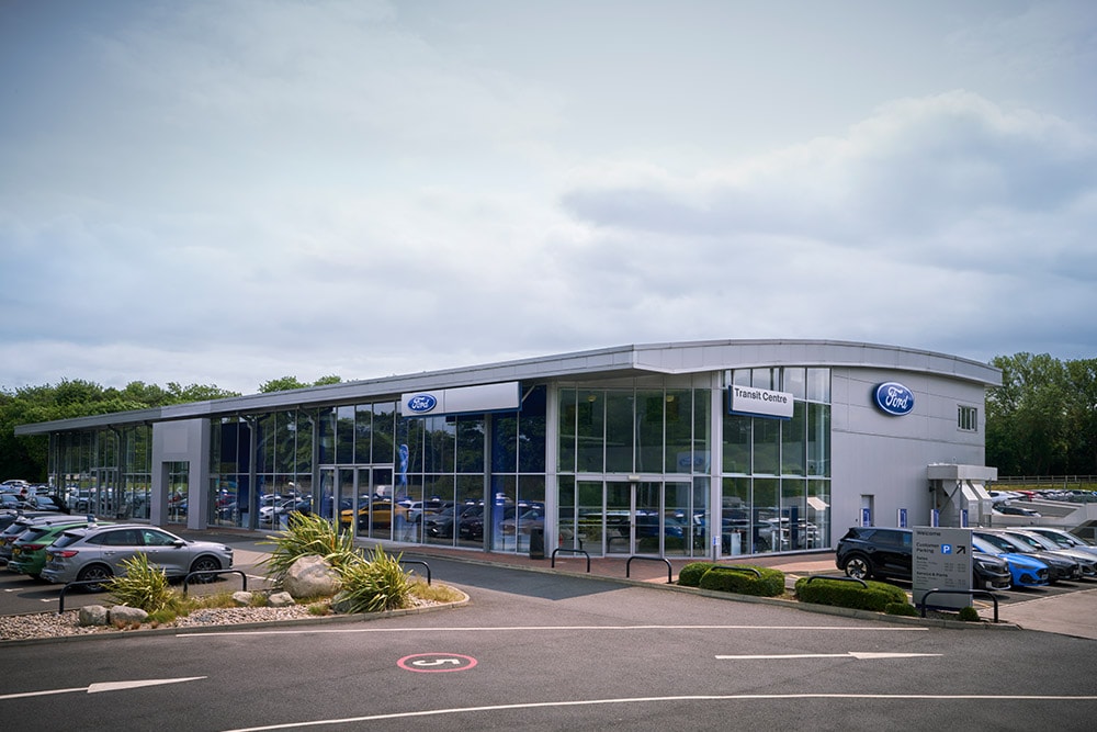 Außenansicht eines modernen Ford-Autohauses mit verschiedenen Ford-Fahrzeugen davor.