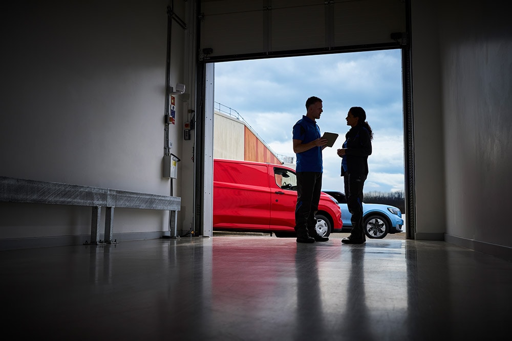Zwei Personen unterhalten sich in einer Garageneinfahrt, vor der ein roter Ford-Transporter und ein blauer Ford-SUV geparkt sind.