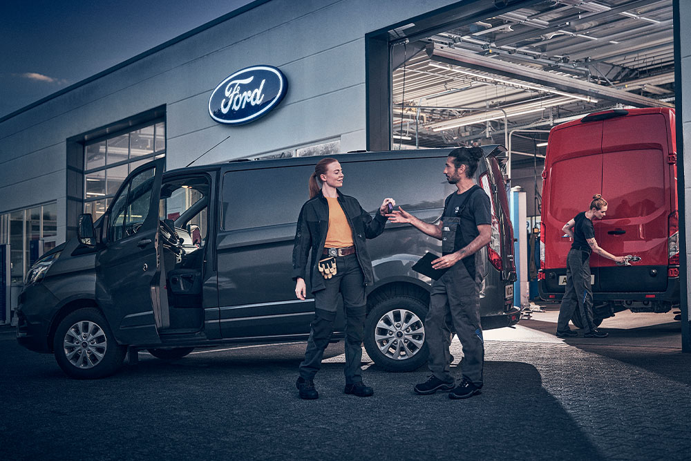 Kunde und Ford-Techniker tauschen Schlüssel für einen Transporter vor einem Ford-Autohaus aus.