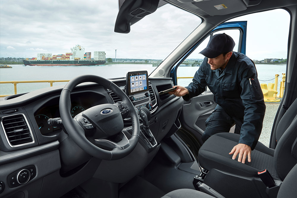 Ein Mann, der sich in die offene Tür eines Ford-Vans lehnt und ein Telefon in Händen hält, während ein Hafen im Hintergrund zu sehen ist.