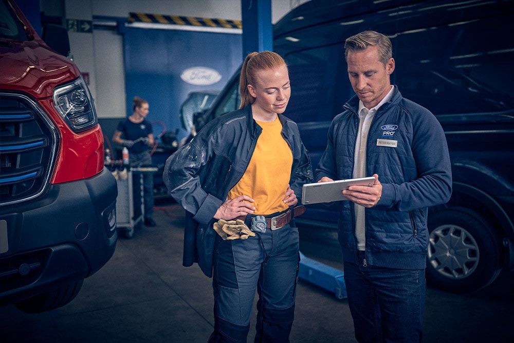 Ein Mann und eine Frau in Arbeitskleidung betrachten ein Tablet in einer Garage mit einem roten Ford-Van.