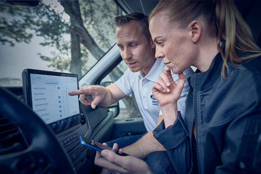 Ein Mann zeigt auf einen großen Touchscreen in einem Van, eine Frau schaut zu und hält ein Smartphone in der Hand.