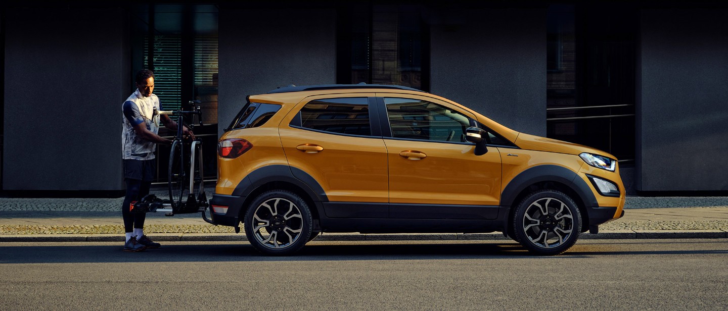 Ford EcoSport in orange Seitenansicht parkend auf Straße in Stadt während ein Mann ein Fahrrad auf dem Heckträger befestigt.