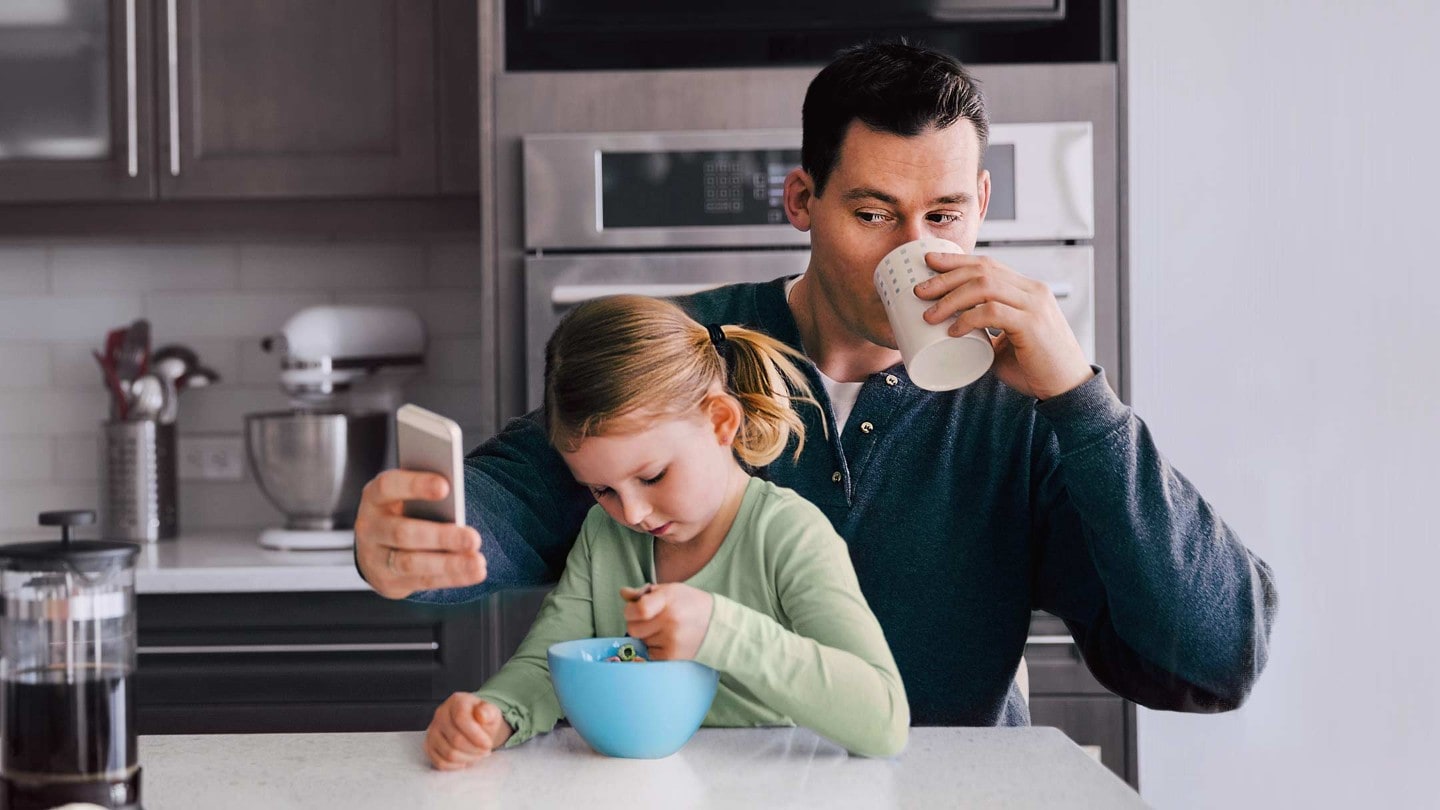 Mann mit Kind auf dem Schoss trinkt aus einer Tasse und schaut auf sein Smartphone