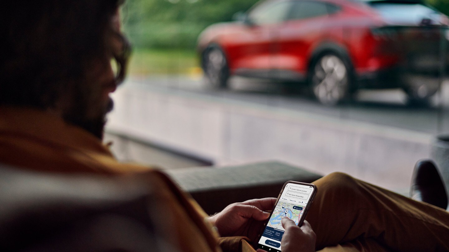 Sitzender Mann schaut auf sein Smartphone, mit einem Ford Mustang Mach-E in Rot im Hintergrund