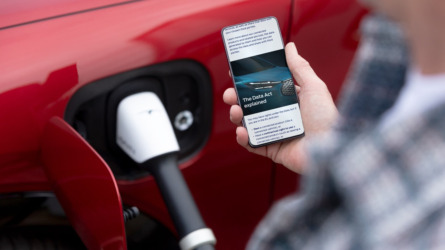 Eine Person checkt ihr Smartphone, ein rotes Elektrofahrzeug lädt im Hintergrund auf.