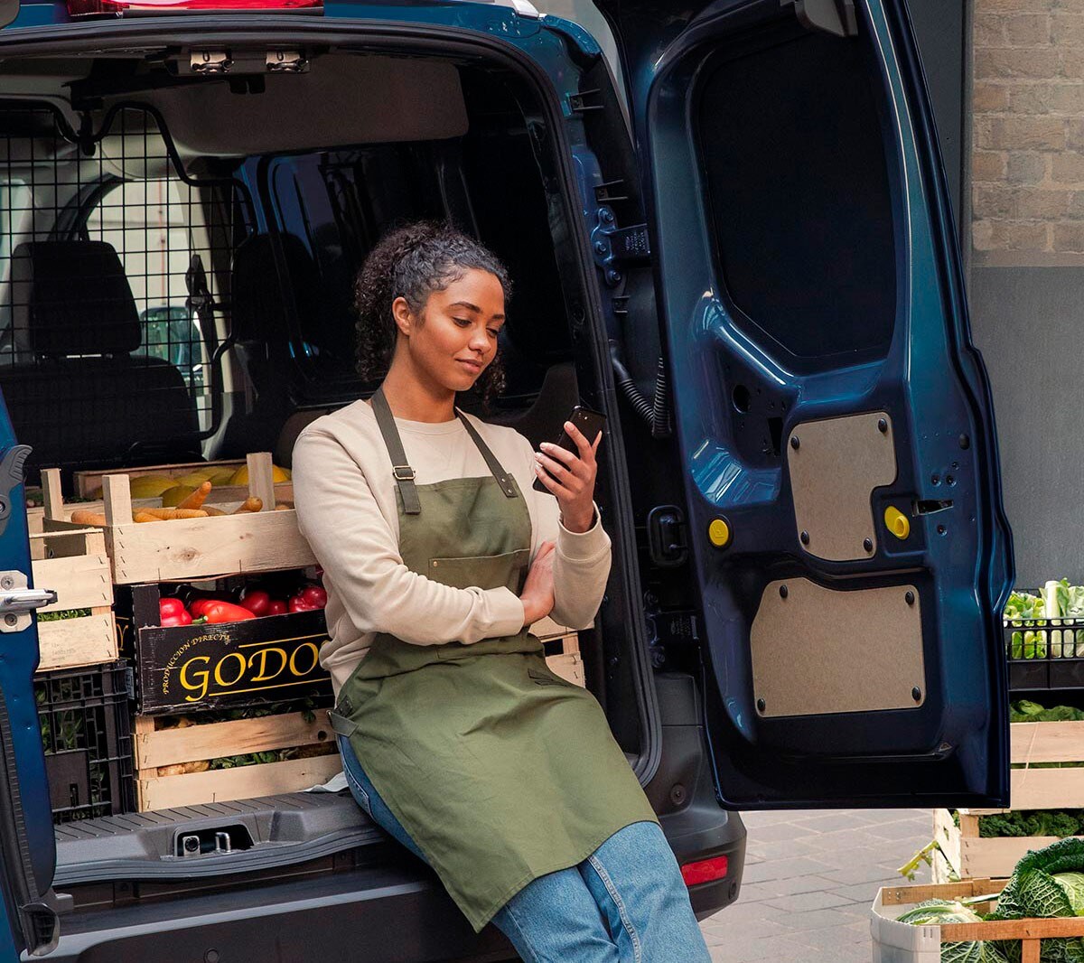 Händlerin schaut auf ihr Handy und lehnt an der Lädeflache eines Ford Transit Couriers.