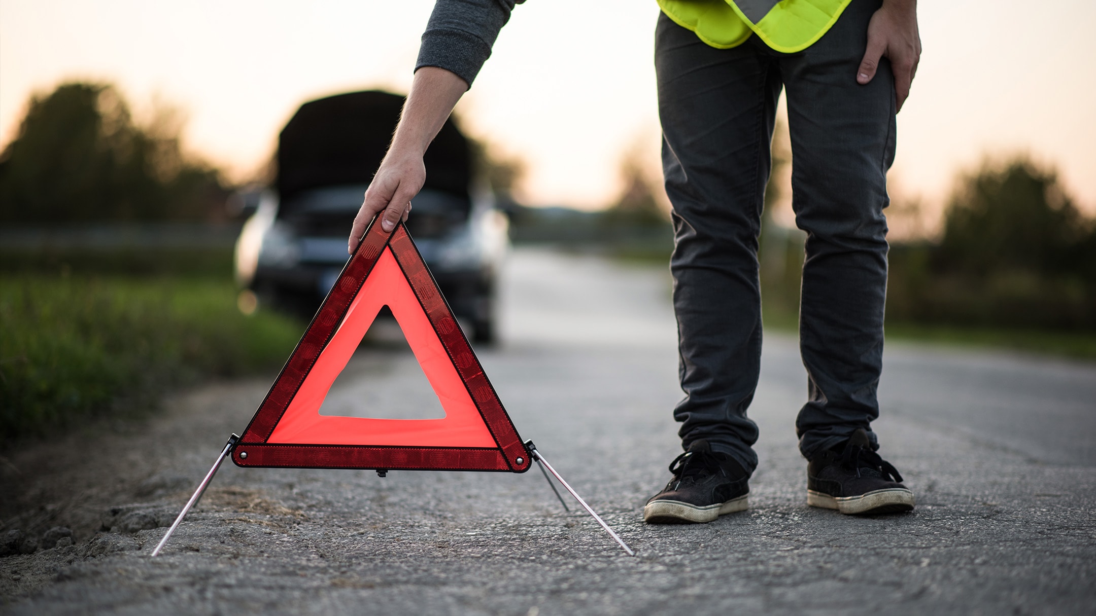 Eine Person stellt ein rotes Warndreieck auf, da ein Fahrzeug am Straßenrand liegen geblieben ist.