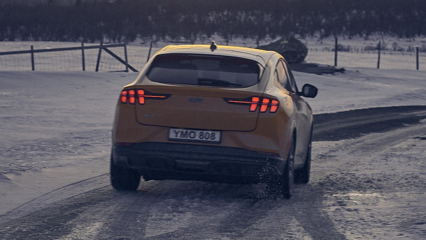 Ford Mustang Mach-E in orange fahrend auf Schnee bedeckter Straße in den Bergen. Heckansicht. 