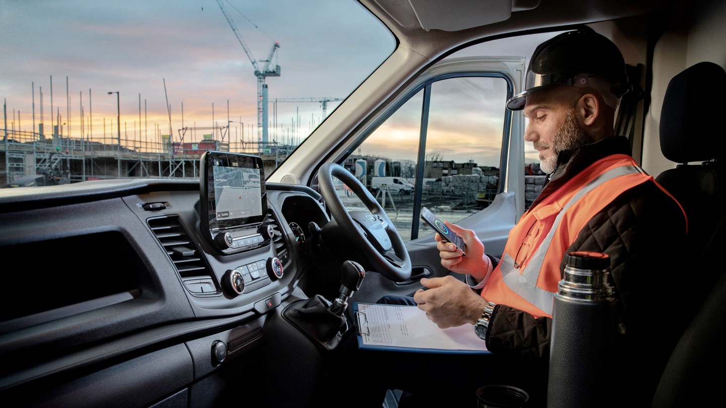 Arbeiter mit Bauhelm sitzt hinter dem Steuer eines Fahrzeugs und schaut auf ein mobiles Telefon