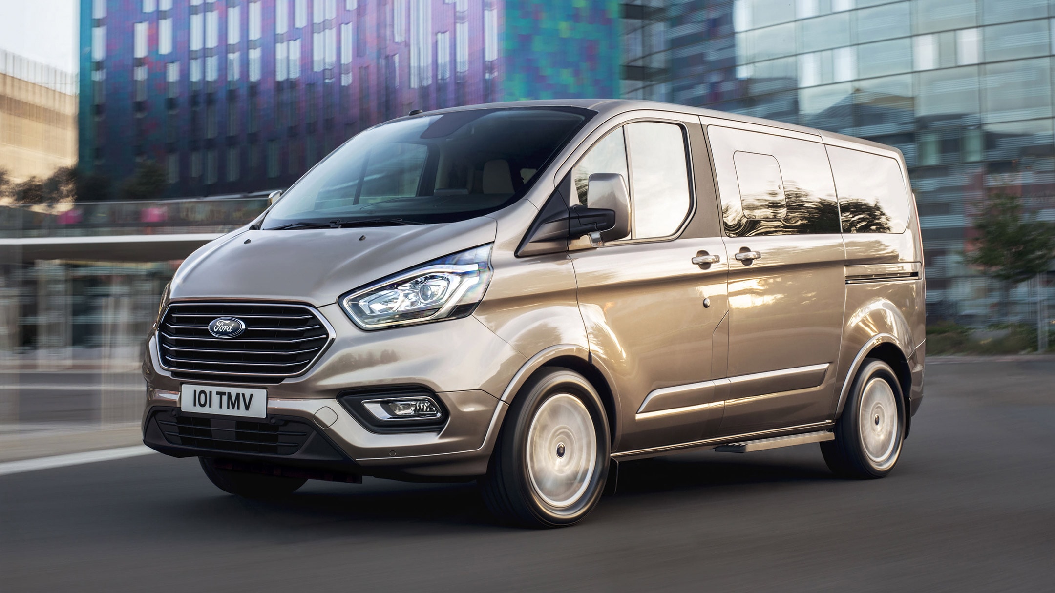 Ford Tourneo  Custom Champagner ¾-Frontansicht in Bewegung vor modernem Gebäude